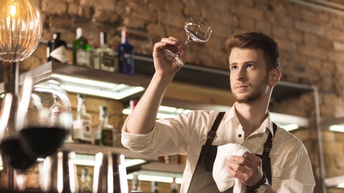 Person mit kurzen braunen Haaren und Bart steht hinter einer Bar und poliert ein Weinglas