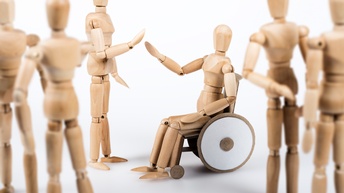 Drei Holzfiguren und eine der Figuren sitzt in einem Rollstuhl