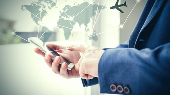 Seitliche Aufnahme der Hände einer Person, die ein Smartphone hält. Die Person trägt ein blaues Sakko. Darüber ist eine Grafik einer Weltkarte gelegt. Neben der Weltkarte ist ein Flugzeug