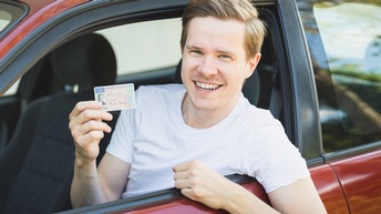 Lächelnde Person lehnt sich auf Fahrerseite aus Auto und hält Führerschein in Hand