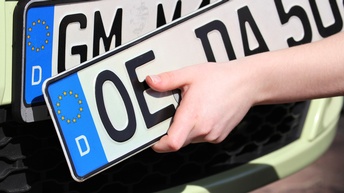 Hand hält ein weiteres Autokennzeichen zu einem Fahrzeug mit montiertem Autokennzeichen