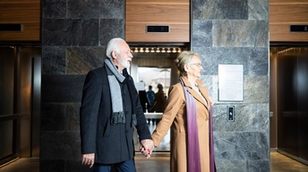 Zwei Personen mit Schals in Seitenansicht hintereinander Hand in Hand gehend, im Hintergrund Lifte mit geöffneten Türen