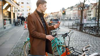 Person steht auf Nebenstraße, hält eine Hand auf Fahrrad und in der anderen Smartphone, im Hintergrund Stadtgeschehen