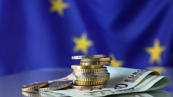 Verschiedene Euromünzen liegen gestapelt. aneinandergereiht auf mehreren Euroscheinmünzen, im Hintergrund zeigt sich eine blaue Flagge der europäischen Union mit einem Ausschnitt von gelben Sternen