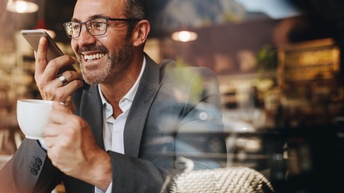Lächelnde Person in Anzug mit breitem Silberring hält Smartphone zum Mund und in anderer Hand Kaffeetasse