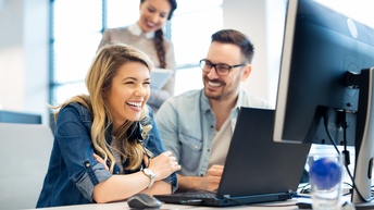 Zwei Personen vor Computerbildschirm sitzend lachen, im Hintergrund verschwommen weitere lachende Person