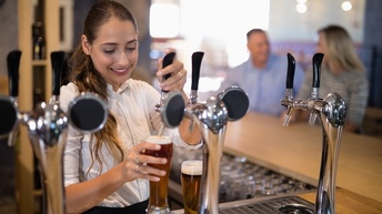 Lächelnde Person zapft Bier an einer Theke in Glas, im Hintergrund zwei weitere Personen in Unterhaltung miteinander