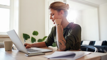 Person mit zusammengebundenen blond-braunen Haaren und Tattoos auf den Händen sitzt mit einem grünen Blusenkleid an einem Schreibtisch und notiert sich etwas während daneben ein Laptop, ein Smartphone sowie Unterlagen liegen