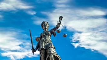 Statue der Justitia, Schwert und Waagschale haltend, im Hintergrund blauer Himmel durchzogen von Wolken