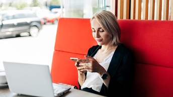 Person mit blonden Haaren sitzt  bei einem Tisch in einem Restaurant und blickt auf ein Smartphone während ein Laptop und eine Tasse auf dem Tisch stehen