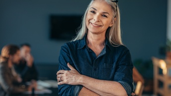 Person mittleren Alters mit langen weiß-grauen Haaren, Brille und blauem Jeanshemd steht lächelnd und mit verschränkten Armen in einem Raum während im Hintergrund andere Personen sitzen