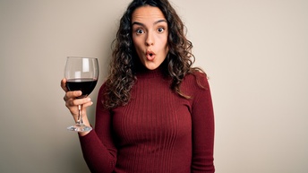 Eine Person in einem dunkelroten Rollkragenpullover, mit langen, dunkelbraunen Locken steht vor hellgrauem Hintergrund. In der rechten Hand hält sie ein Glas Wein befüllt mit Rotwein. Ihr Mund ist zu einem O geformt