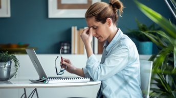 Eine Person sitzt an einem Schreibtisch mit Arbeitsunterlagen und aufgeklappten Laptop und greift sich mit der Hand zwischen die geschlossenen Augen.