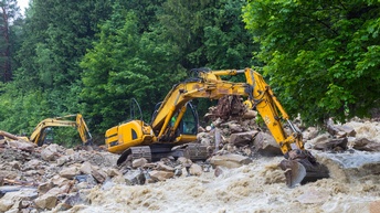 Zwei gelbe Bagger auf steinigem Geröll stehend schaufeln in reißendes Wasser, im Hintergrund entwurzelter Baum und grüner Wald