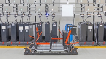 Reihe grauer Industriebatterien verbunden mit Kabeln zu grauen Boxen, im Vordergrund Hebegerät
