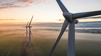 In der rechten Bildhälfte ist die Nahaufnahme des oberen Teils eines Windrads mit drei langen Metallstäben. Im Hintergrund sind zwei weitere Windräder. Alle drei stehen auf einer grünen, nebelverhangenen Wiese.