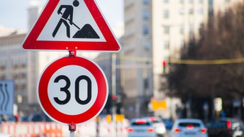 Zwei Straßenschilder im Fokus: Ein Baustellen- und ein 30-er-Zonene-Schild, im Hintergrund verschwommen Autos in Rückansicht.