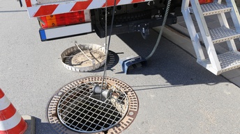 Neben einem Kanaldeckel steht ein Fahrzeug mit einer Trittbrettleiter hinten dran. Vom Fahrzeug hängen zwei Schnüre in den Kanaldeckel hinein