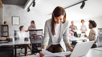Person mit langen dunklen Haaren und Businesskleidung steht vor einem Bürotisch und blickt dabei auf Unterlagen während sie einen Laptop bedient, im Hintergrund sitzen und stehen weitere Personen rund um Schreibtische in einem Büroraum