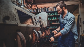 Bärtige Person mit Brillen in Arbeitsschürze steht an großer Poliermaschine und hält Lederschuh an Riemen, im Hintergrund Regale mit Spraydosen und Schuhmodeln