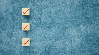 Es ist eine Checkliste dargestellt. Auf einem türkis-blauen Polster-Hintergrund aus Samt sieht man im linken Bildteil drei quadratische Holzwürfel, auf denen rote Häkchen abgebildet sind. Das Bild wirkt aufgeräumt und hat Struktur. <br />
