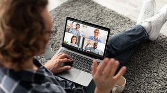 Eine Person sitzt am Boden auf einem Teppich. Auf dem Schoß hat sie einen Laptop, auf dessen Bildschirm vier Personen sind. Neben der Person sind ein Smartphone und eine Tasse