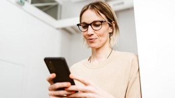 Nahaufnahme einer Person mit Brille, die lächelt, sodass sich Grübchen neben dem Mund bilden. Ihr Blick ist auf ein Smartphone gerichtet, das sie in beiden Händen hält. Sie steht in einem sterilen Raum 