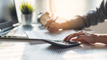 Detailansicht auf Hand einer Person, die an Schreibtisch sitzt und auf Taschenrechner tippt, ringsum Arbeitsdokumente und aufgeklappter Laptop