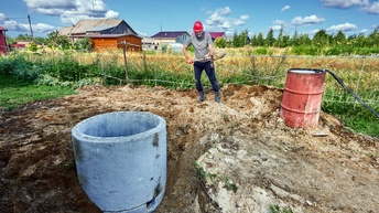 Eine Person schaufelt Sand, er steht neben einer Grube, aus der ein Brunnen ausgehoben wird. Neben ihm ist eine Tonne aus Metall. Im Hintergrund eine Landschaft mit Häusern, es herrscht Sonnenschein. 