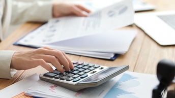 An einem Tisch sitzt eine Person in einem hellen Sakko. Die Person tippt mit der rechten Hand auf einem Taschenrechner. Darunter sind mehrere Zettel mit verschiedenen Diagrammen