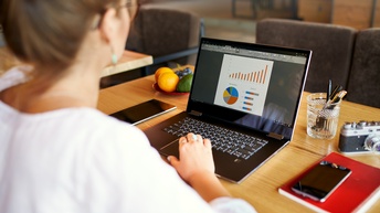 Person sitzt vor einem Laptop in einem Restaurant und betrachtet Grafiken, Diagramme sowie liegen Stifte, Notizbücher, eine Kamera und ein Smartphone, ein Tablet und eine Schale Obst