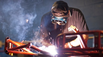 Person in Schutzbekleidung mit Schutzbrille und -Handschuhen führt Schweißerarbeiten durch und Funken erleuchten den Raum