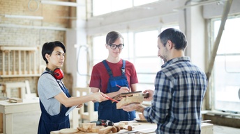 Drei Personen stehen um einen Werktisch in einer großen Werkstatt. Eine der Personen hält ein Holzstück und zeigt es den anderen beiden. Die anderen Personen halten jeweils einen Block und einen Stift