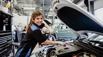 Seitliche Aufnahme einer Person mit Handschuhen, die an einem Auto mit geöffneter Motorhaube steht und den Ölstand misst. Die Person blickt direkt in die Kamera. Im Hintergrund stehen weitere Autos