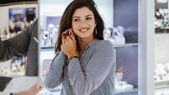 Eine Person steht in einem Geschäft und gibt sich einen Ohrring in das rechte Ohr. Im linken Ohr ist bereits ein Ohrring. Eine Person hält ihr einen Spiegel hin, in dem sie sich betrachten kann