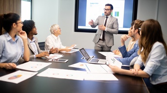 Personen in Businesskleidung sitzen an einem Besprechungstisch mit Unterlagen, Tablet und Laptop, eine Person mit kurzen Haaren und Brille im Anzug steht vorne und präsentiert, im Hintergrund wird eine Präsentation angezeigt