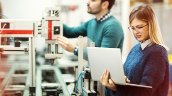 Person mit Brille in einer technischen Produktion hält einen Laptop in der Hand und blickt darauf, zweite Person in Unschärfe bedient eine Maschine im Hintergrund