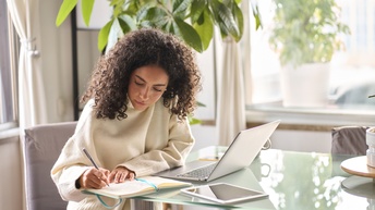 Eine Person sitzt seitlich an einem Glastisch. Vor der Person ist ein aufgeklappter Laptop. Rechts daneben ist ein aufgeklapptes Notizbuch, in das die Person etwas schreibt. Dahinter liegt ein Tablet. Im Hintergrund sind Topfpflanzen und Fenster