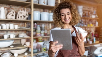Person mit lockigem Haar und roter Arbeitsschürze hält eine Tasse und blickt freudig auf ein Tablet, im Hintergrund zeigt sich ein Regal mit Tonwaren und Arbeitsmaterialien