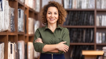 Person mit lockigen, schulterlangen Haaren und  grüner Bluse steht mit verschränkten Armen freudig vor Regalen mit Bettwaren