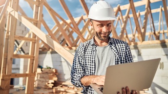 Eine Person mit Helm hält einen aufgeklappten Laptop und blickt darauf. Im Hintergrund ist eine Mauer aus grauen Ziegeln. Darauf ist ein Dachstuhl aus Holz. Unter dem Dachstuhl liegen viele Holzbalken gestapelt und verstreut