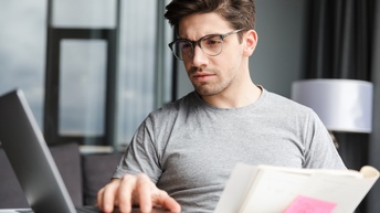 Eine Person mit Brille an einem Tisch. Vor der Person steht ein geöffneter Laptop, den sie mit einer Hand bedient. Rechts daneben liegt ein geöffneter Notizblock mit einem pinkfarbenen Klebezettel. Die Person blickt auf den Bildschirm des Laptops