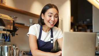 Person in Arbeitskleidung mit blauer Schürze steht in einem Café an der Theke und blickt freudig in einen Laptop