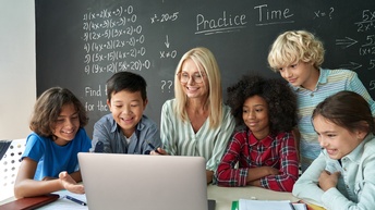 Lehrperson sitzt mit Schüler:innen vor aufgeklappten Laptop, im Hintergrund beschriebene Kreidetafel