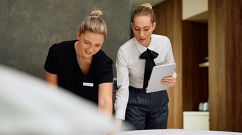 Eine Hausdame in einem Hotel zeigt einem Zimmermädchen das Bettenmachen