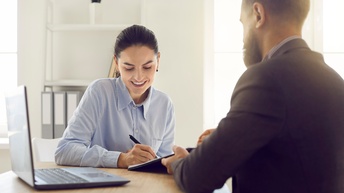 Zwei Personen sitzen sich an Tisch gegenüber, eine Person hält Dokument, das andere Person unterfertigt, am Tisch aufgeklappter Laptop