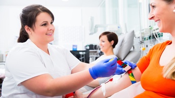 Seitliche Aufnahme zweier sich gegenüber sitzender Personen, die sich anblicken. Die linke Person trägt Handschuhe und hält ein buntes Band um den Oberarm der rechten Person. Aus dem Oberarm kommen schmale Schläuche mit Blut.