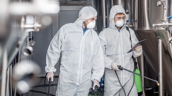 Zwei Personen in weißem Schutzanzug mit Brille und FFP-Maske führen Reinigungsarbeiten in einer Brauerei durch und blicken dabei auf ein Tablet