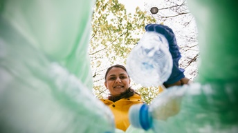 Person mit dunklen geschlossenen Haaren trägt Pet-Flaschen zusammen, Blick in die Kamera zwischen den Pet-Flaschen hindurch