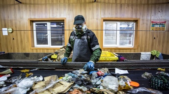 Person mit Kappe, Brille, Handschuhen, in Schutzkleidung und mit Atemschutzmaske steht an Fließband mit Abfällen und greift danach, im Hintergrund Holzwand mit Fenstern, davor stehen Abfalltonnen.